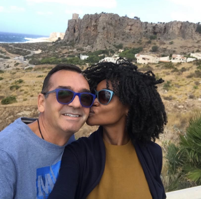 A couple smiling with the view of a rock and the sea behind them