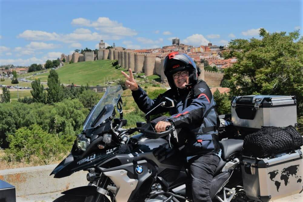 A motorcyclist on his motorcycle with 2 fingers up in a wave. There is the backdrop of the medieval walled city of Avila, Spain behind him.