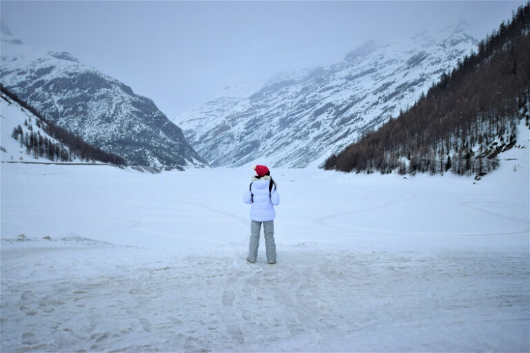 A woman standing and looking at a frozen lake with the snow-covered mountains on both sides in Livigno- A low-budget destination in Italy