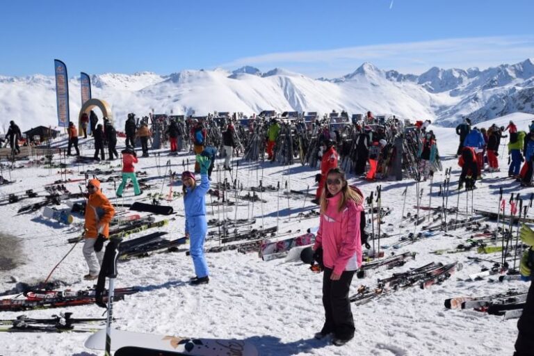 An outdoor winter scene on a ski slope with many skis on the grounds and a few people smiling at the camera in Livigno, a low-budget winter destination in Italy