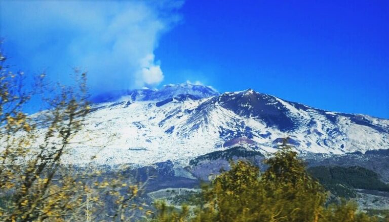 A snow-capped Mt. Etna, erupting smoke from the volcano mouth, with some greenery around. Represents a low-budget destination in Italy