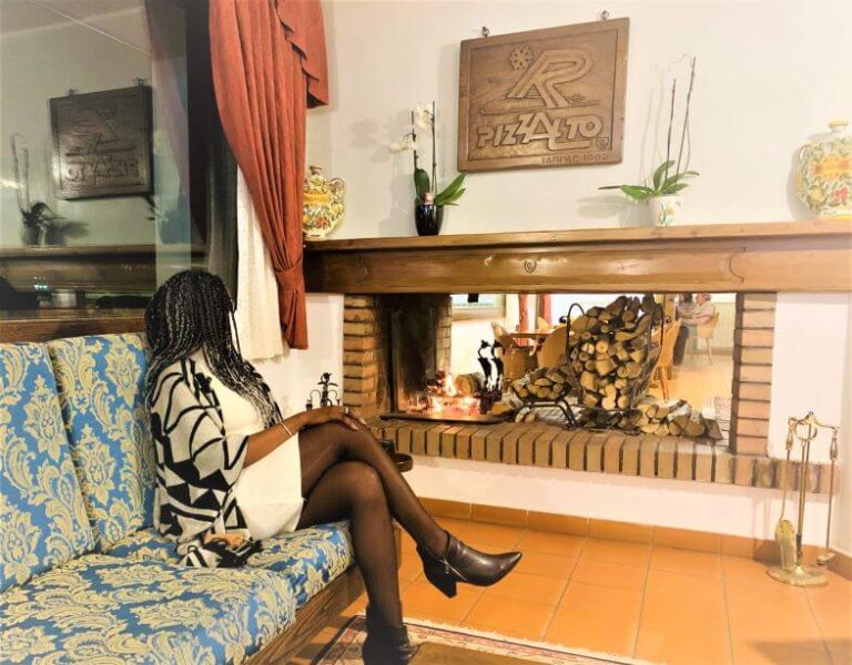 A woman sitting next to a wood fireplace. It represents a low-budget winter destination in Italy