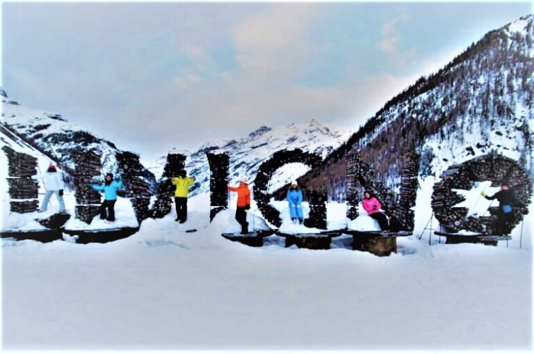 6 people standing next to a sign "LIVIGNO", with the backdrop of the snow-capped mountains. It represents a low-budget winter destination in Italy