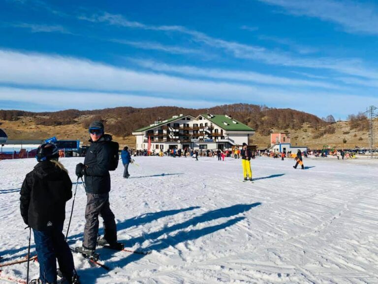 People standing in front of a lone mountain lodge with snow on the ground. Roccaraso is a low-budget winter destination in Italy