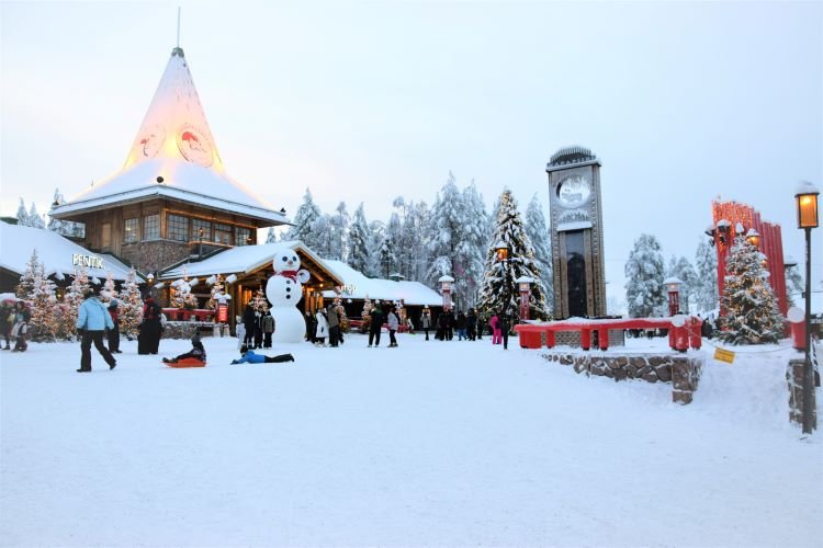A winter scene in a square with a tall clock on the right, a building with a high lighted dome in the center and people walking around a giant snow man in Rovaniemi Lapland