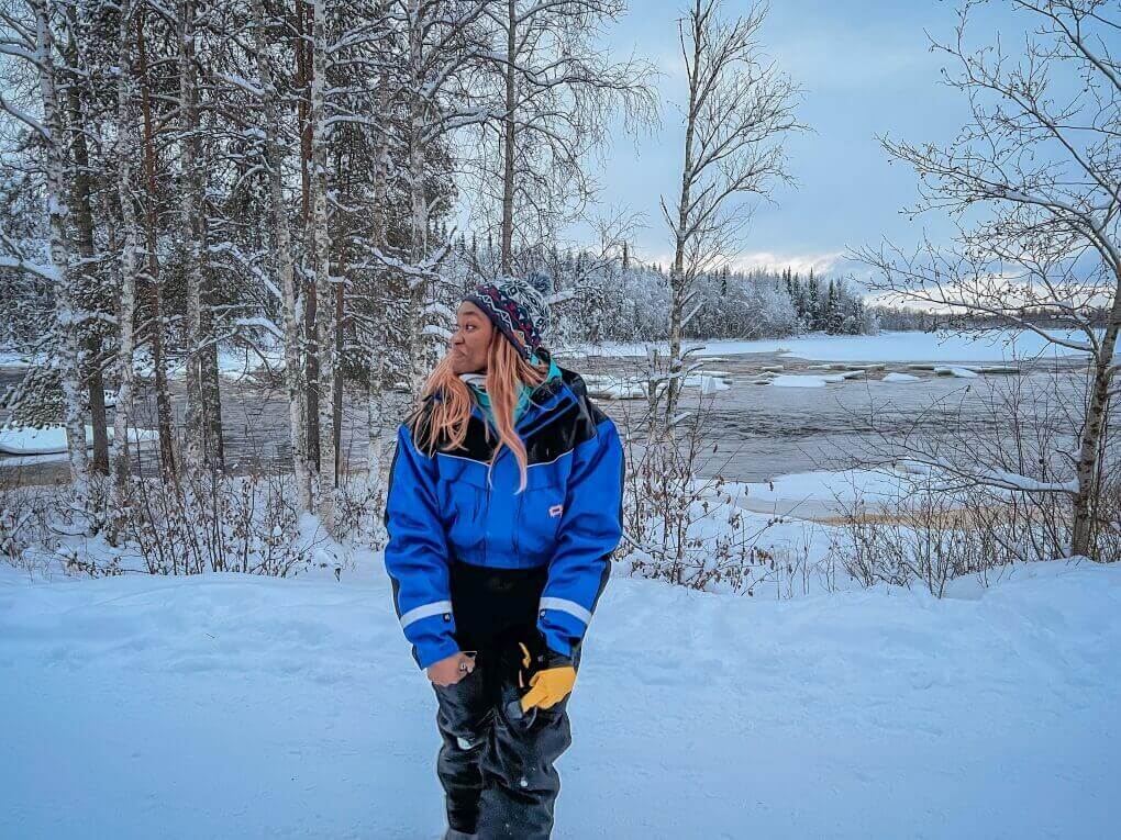 A woman in winter overalls, hat, and gloves standing next to an icy river surrounded by trees in Rovaniemi, Finland