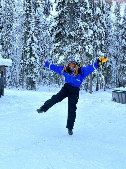 A woman in warm winter overalls jumping with arms outstretched and one leg up. She is outdoors in a winter environment in Rovaniemi, Finland.