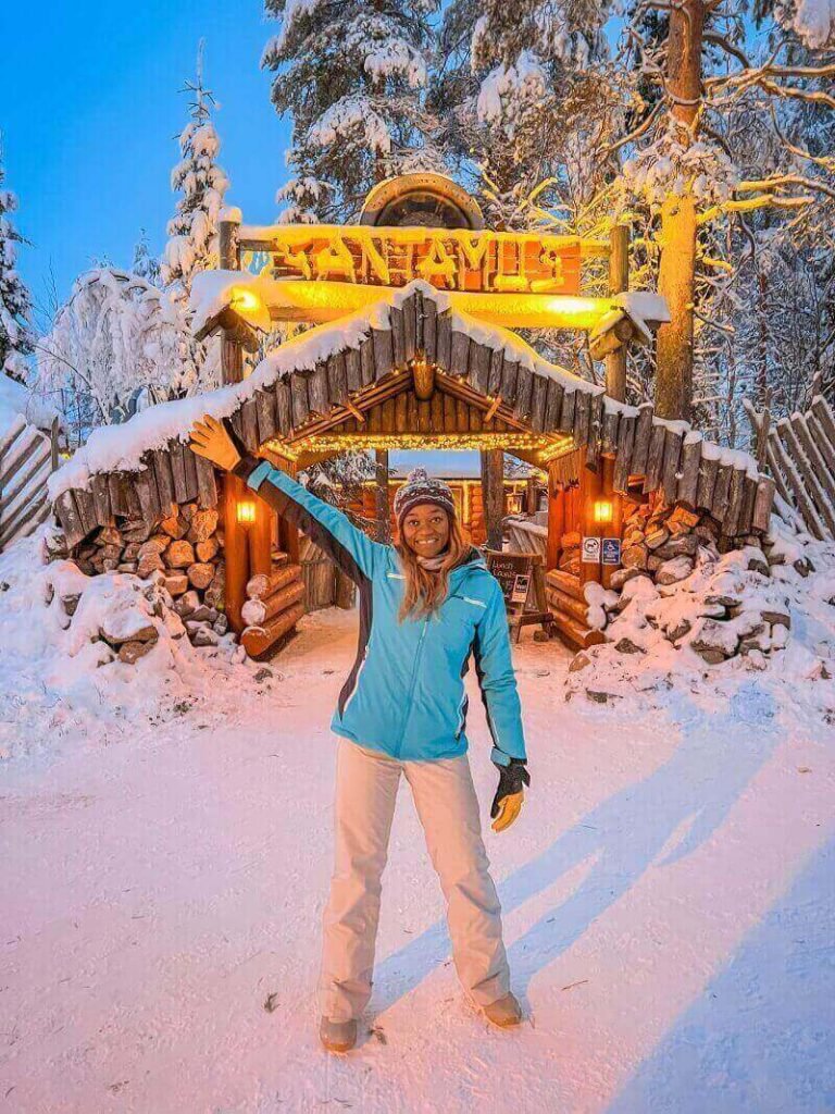 A woman in front os an arctic log cabin with her right arm raised in Santa Claus Village