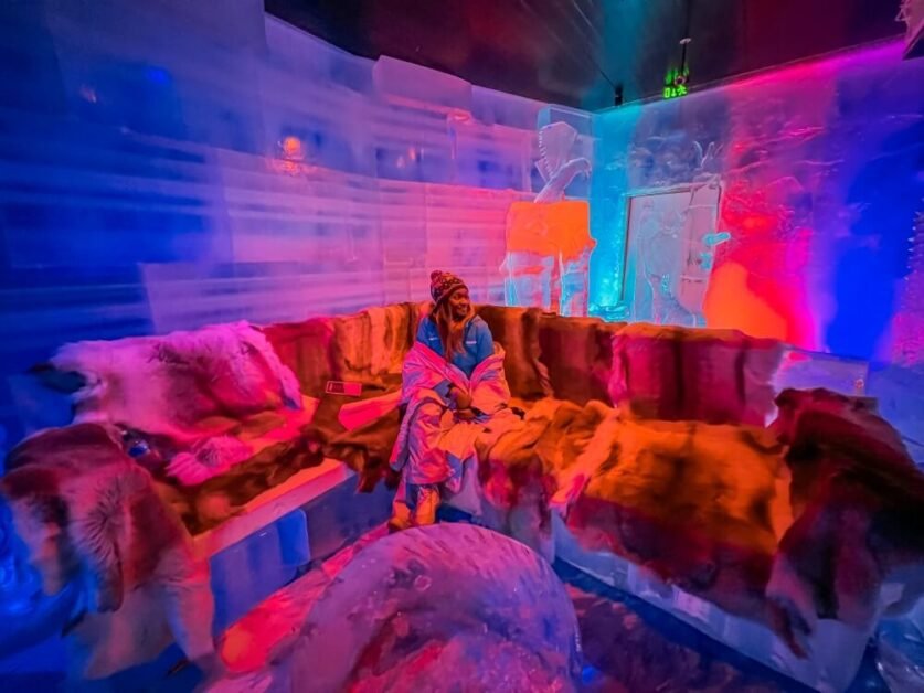 A woman sitting on a long semicircle-shaped divan made out of ice. There are reindeer furs to provide warmth in SantaPark, Rovaniemi