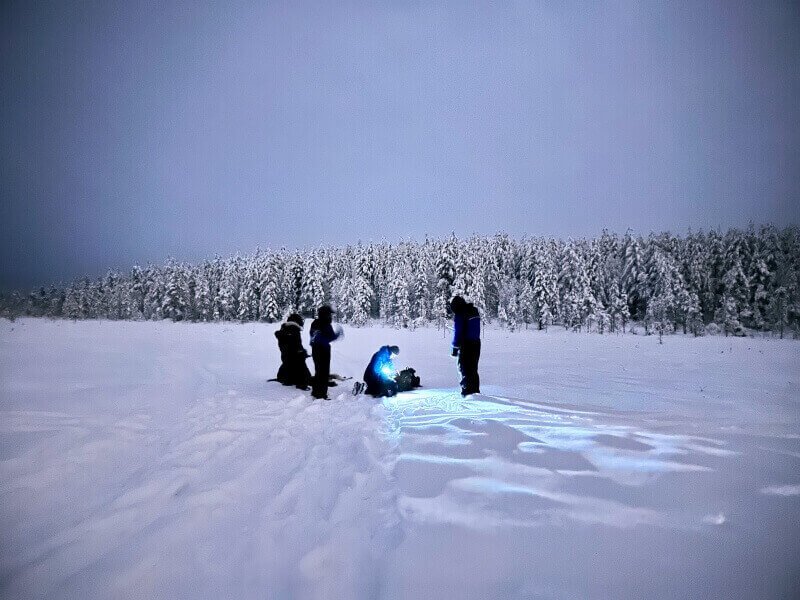 Three people standing around a person who is crouching with a torch light. They are in a winter landscape