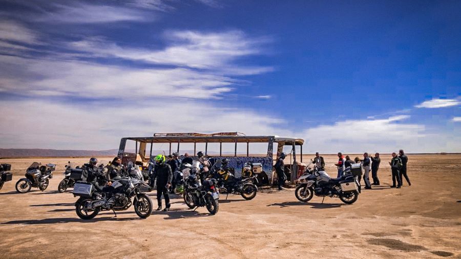 A group of motorcyclists parked and standing in front of a run-down dilapidated bus. They are standing on the base of a dried-up lake called a chott, in Tunisia