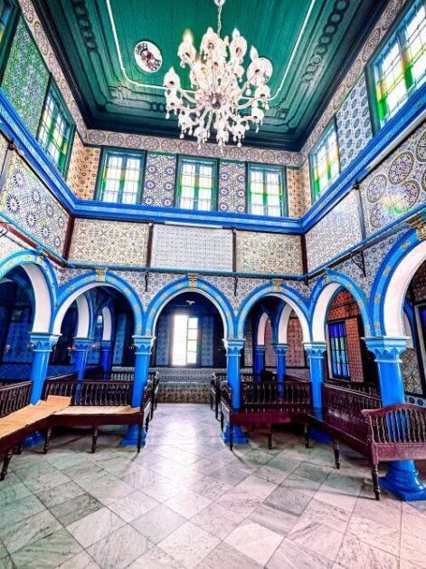 The inside hall of a synagogue. there are several arches and benches. The walls and ceiling are decorated with blue and green tiles