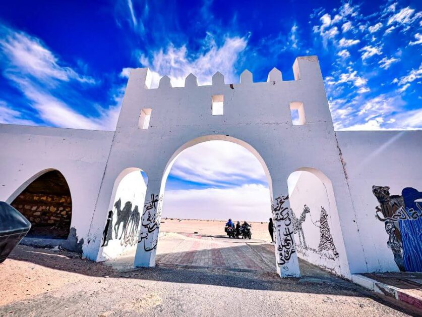 A high, white archway leading to the Sahara Desert. There are some motorcyclists on a motorcycle on the other side of the archway