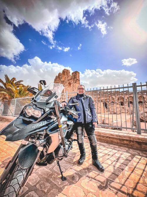 A man standing next to a black motorcycle. The are in front of a gated area. Behind the gate there is a partial view of a roman amphitheatre in Tunisia