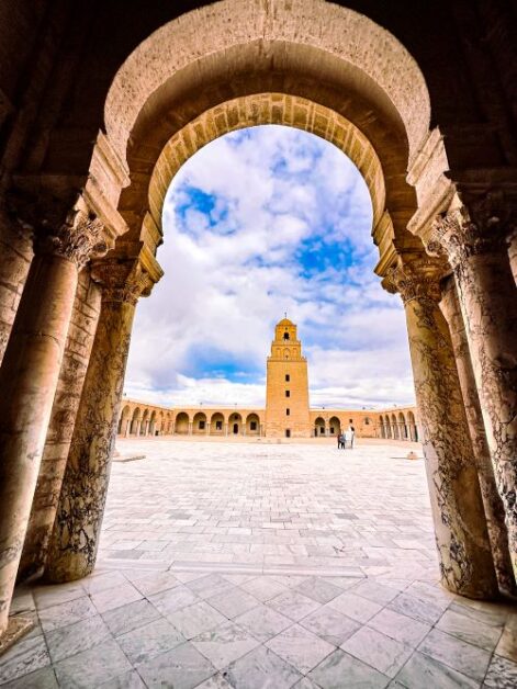 A view from the arches of a building looking out towards a courtyard. There is a another building on the other end with a tower in the center