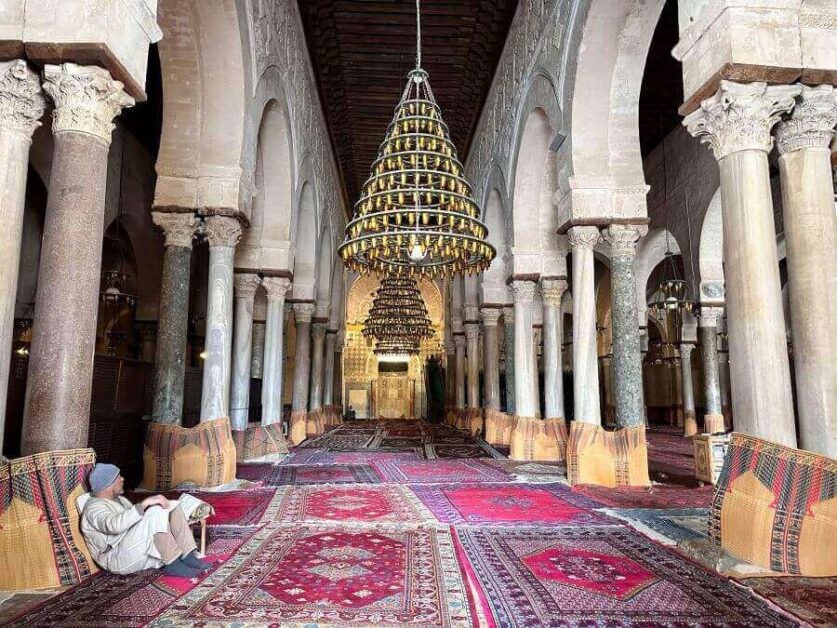 The inside of a mosque with high ceilings, red rugs, giant chandeliers and a forest of columns