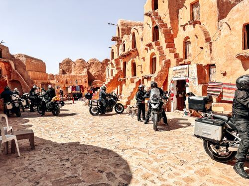 A group of motorcyclists in a courtyard of a structure made of mud, three storeys high, with stairs in front of them.