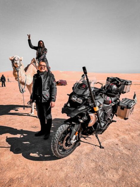 A man standing next to a black motorcycle. Behind him, a woman is sitting on a camel with her right hand raised. They are in a desert landscape