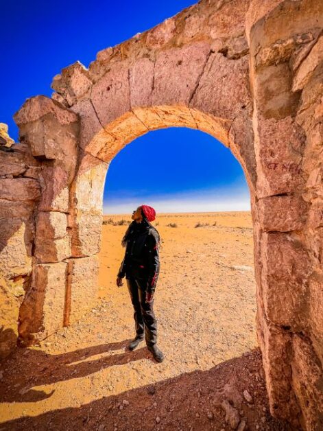 A woman standing under the ruins of an arch and staring upwards. The landscape is in a desert