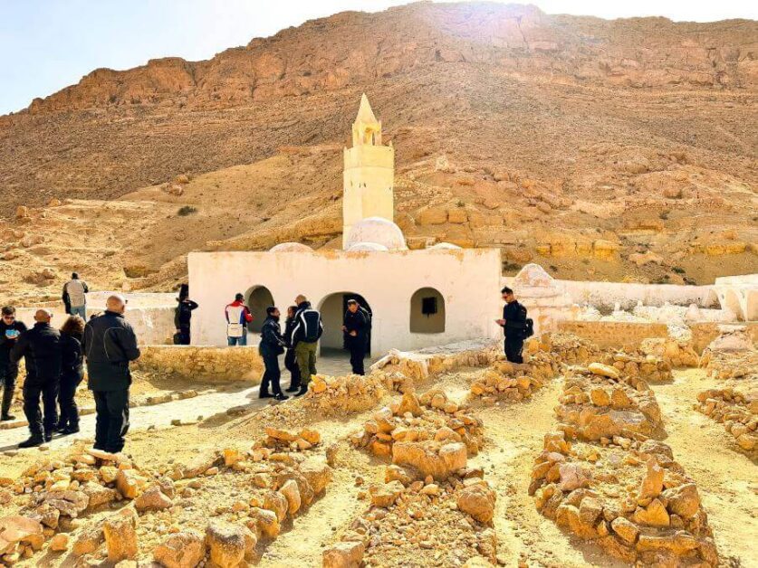People standing infront of a white mosque. The mosque is in a mountain region. There are graves made of stones in front of the mosque. There is a hill behind the mosque