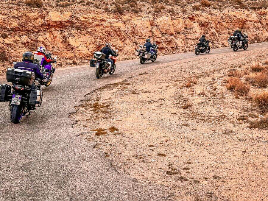 A group of motorcyclists riding up a winding road up the side of a mountain. The landscape is reddish brown in colour