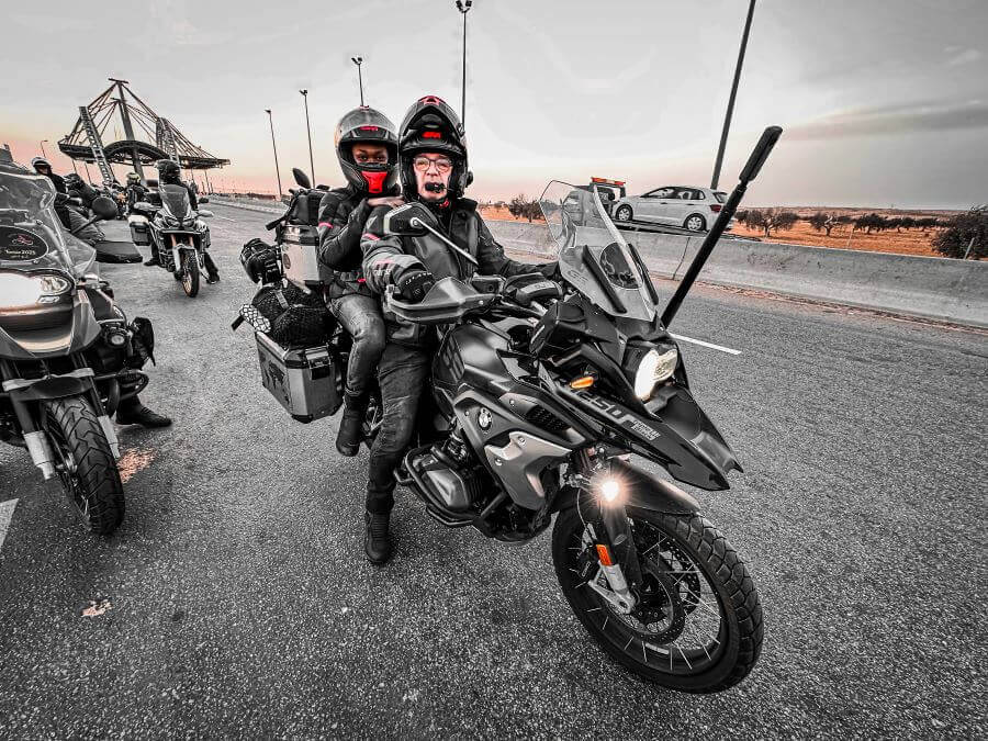 A couple in motorcycle gear on a black motorcycle. They are stopped on the road and looking at the camera