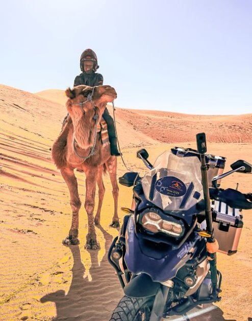 A female motorcyclist on a camel in the desert of Tunisia. There is a black motorcycle parked next to the camel