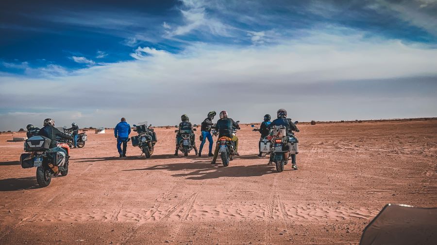 A group of motorcyclists riding in a desert landscape.The view is from behind. Some are on their motorcycles and 3 motorcyclists are on foot