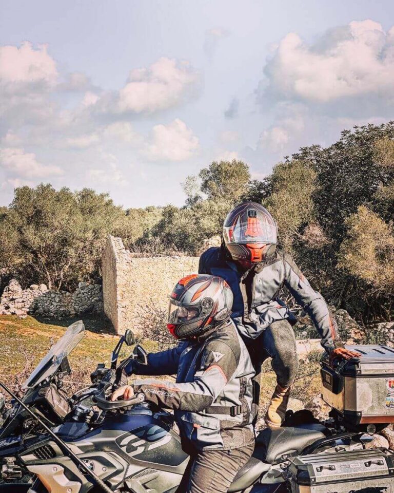 A motorcycle couple outdoors. The passenger is in the process of mounting the motorcycle