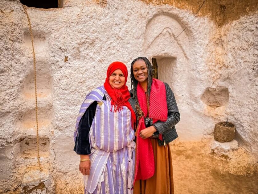 Two women standing next to each other and smiling at the camera. They are in the courtyard of an underground cave home in Matmata
