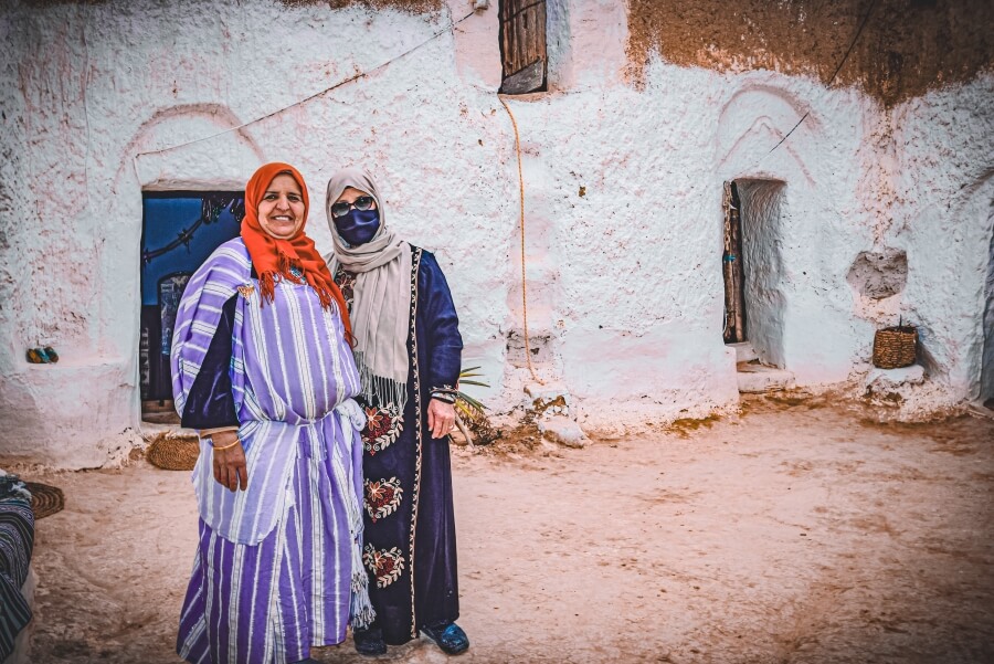 Two women standing next to each other and smiling at the camera. They are in the courtyard of an underground cave home in Matmata. The woman on the right is wearing a face mask and glasses.