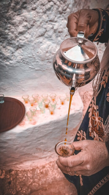 View of the hands a person pouring tea from a silver teapot into a small glass