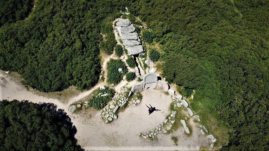 Aerial view of a clearing in a green area with an archeological site shaped like a bull's horn. There is a woman in the center with arms raised and outstretched