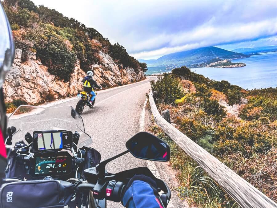 A motorcyclist's view of the curvy road ahead with another rider in front and the view of the sea and vegetations