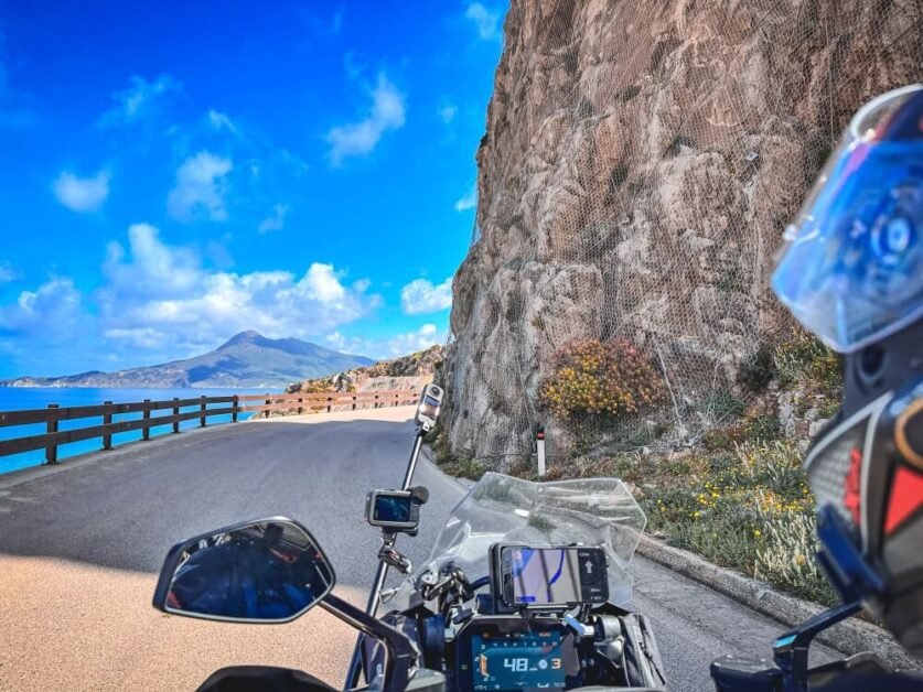 Point of view of a motorcyclist riding on a winding mountain road with a view of the sea and a mountain on the left, in Sardinia
