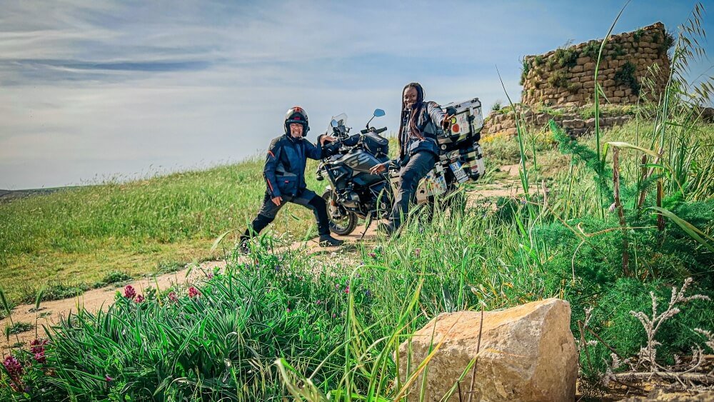 A man and woman dressed in motorcycle gear and standing at each end of a big black motorcycle. They are in a field with the ruins of an old tower to their right