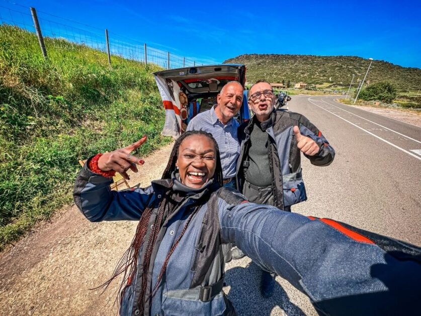 A smiling woman taking a selfie with two men and a van in the background. They are on a countryside road in Sardinia